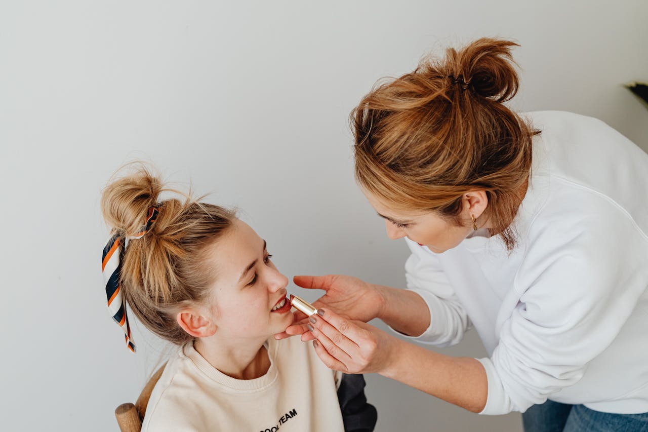 A mother bonding with her daughter by applying lipstick in a cozy indoor setting.