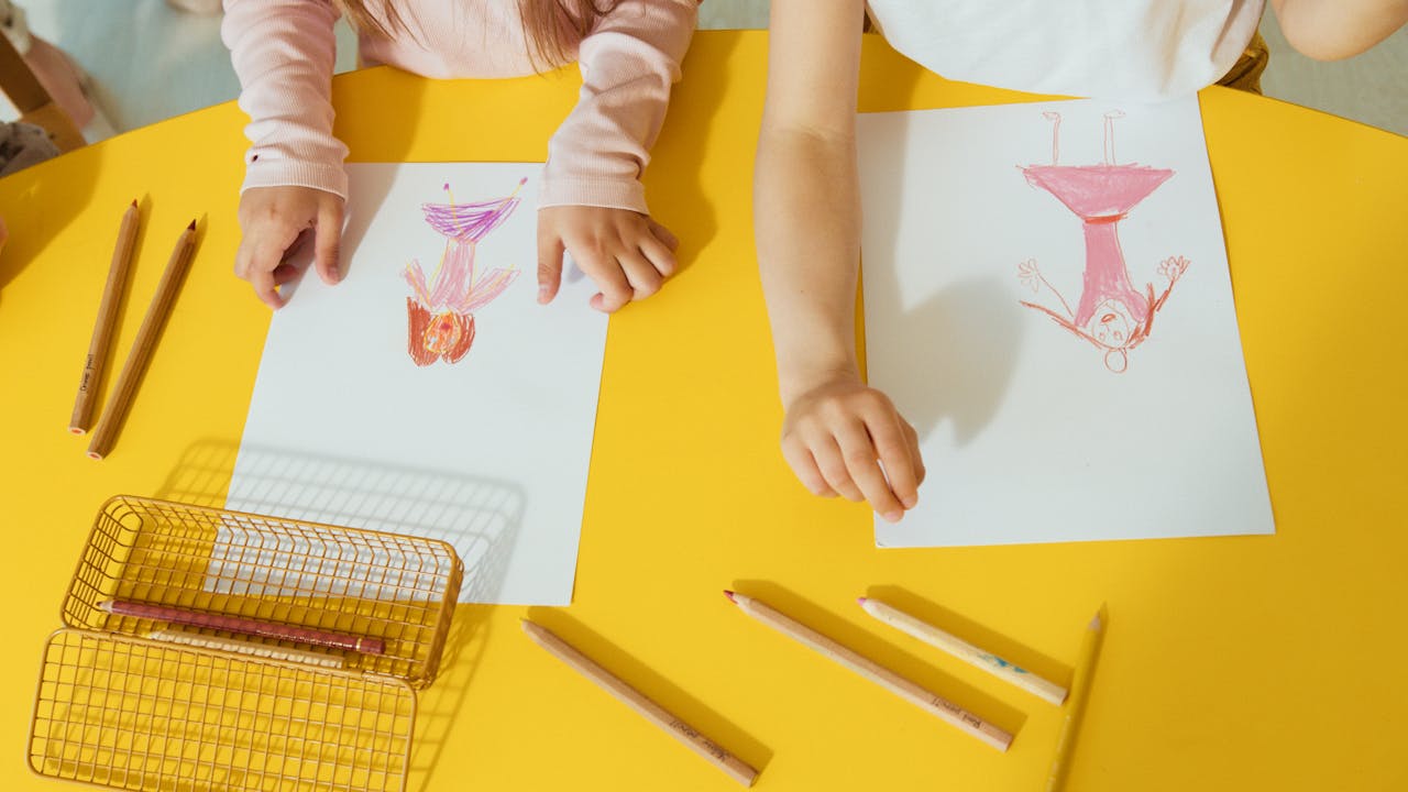 Two children drawing colorful pictures with crayons on a bright yellow table.