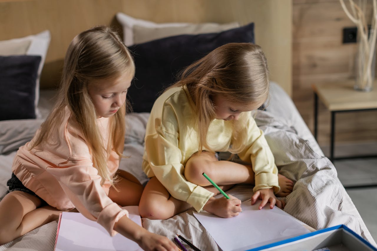 Cute twin girls drawing with pencils while sitting on a cozy bed indoors.